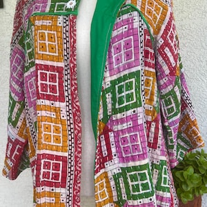 May include: A quilted jacket featuring a vibrant patchwork pattern of squares in yellow, pink, red, and green. The garment has a solid green inner lining, long sleeves, and two white flower-shaped buttons at the top closure with visible white stitching.