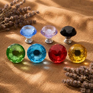 May include: A collection of seven faceted, round cabinet knobs in various colors including blue, light purple, black, green, turquoise, red, and yellow. Each knob features a metallic silver base and a polished, multi-faceted glass top.