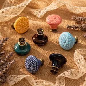 May include: A collection of six ceramic cabinet knobs with brass-toned metal bases. The set includes three knobs with embossed floral patterns in yellow, light blue, and blue, and three smooth, solid-colored knobs in pink, dark brown, and black.