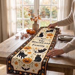 Puede incluir: Un camino de mesa rectangular de tela con un centro color crema decorado con gatos negros, calabazas de Halloween naranjas, hojas de otoño y murciélagos. El texto "The Blackwood Family" está impreso en el centro, con un borde negro y detalles naranjas.