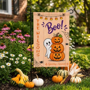 Puede incluir: Una bandera de jardín rectangular con fondo de veta de madera clara que presenta una guirnalda de dulces, el texto morado "Boo!", una pila de tres calabazas de Halloween, un fantasma blanco, una telaraña y el texto naranja "THE WILSONS" y "Spooky Season".
