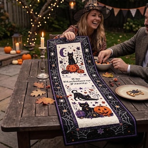 May include: A rectangular quilted table runner featuring a black and white design with purple accents. The fabric displays illustrations of black cats, orange pumpkins, and a crescent moon, with the text The Blackthorne Family printed in the centre.