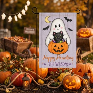 Puede incluir: Una bandera de jardín rectangular con un fantasma blanco que sostiene un gato negro dentro de una calabaza naranja. El fondo es morado claro con una luna creciente amarilla y murciélagos negros. Texto Happy Haunting y THE WILSONS.
