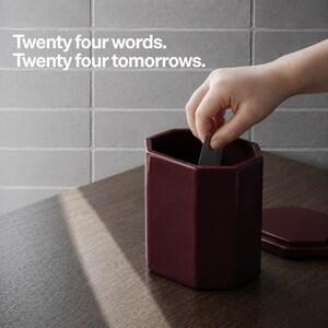 May include: A dark burgundy, octagonal ceramic canister with a matching lid. The text "Twenty four words. Twenty four tomorrows." is printed in white above the container. A hand is shown placing a small, thin, dark rectangular card inside.