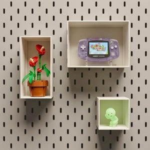 May include: Three white rectangular wall shelves mounted on a light gray pegboard wall. The left shelf holds a red flower plant made of plastic bricks. The top right shelf displays a purple Game Boy Advance console. The bottom shelf holds a small green figure.