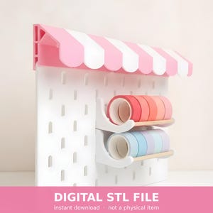 May include: A white pegboard organizer featuring a pink and white striped scalloped awning at the top. Two white shelves are attached to the board, holding rolls of washi tape in shades of red, orange, pink, blue, and purple. Text reads DIGITAL STL FILE.