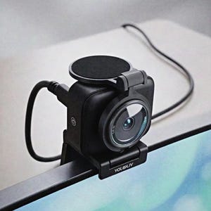 May include: A black YoloLiv webcam mounted on the top edge of a monitor. The device features a circular lens, a power button icon on the side, a black cable connected to the rear, and a circular black cover resting on top of the camera housing.