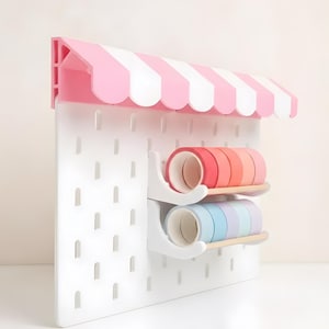 Peut inclure: Un panneau perforé blanc doté d'un auvent rayé rose et blanc sur le dessus. Deux étagères horizontales sont fixées au panneau, supportant des rouleaux de ruban adhésif washi dans des tons de rouge, orange, rose, bleu et violet.