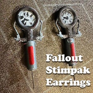 May include: A pair of Fallout Stimpak earrings featuring a circular gauge top with a white face and black markings, connected to a grey cylindrical body with a vertical red rectangular accent and a thin metal needle extending from the bottom.
