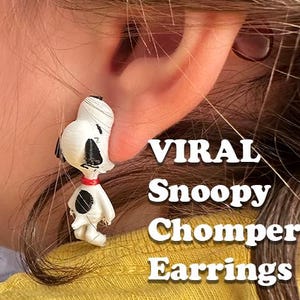 Pode incluir: Um brinco com a forma da personagem Snoopy, em branco e preto com uma coleira vermelha. O texto VIRAL Snoopy Chompers Earrings está impresso a branco no lado direito da imagem, sobreposto ao fundo da fotografia.