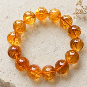 Könnte beinhalten: Ein kreisförmiges Armband bestehend aus vierzehn durchscheinenden, gold-orangen, kugelförmigen Perlen. Jede Perle weist interne Einschlüsse sowie eine polierte, reflektierende Oberfläche auf. Die Perlen sind auf ein dünnes, leuchtend oranges Gummiband gefädelt.