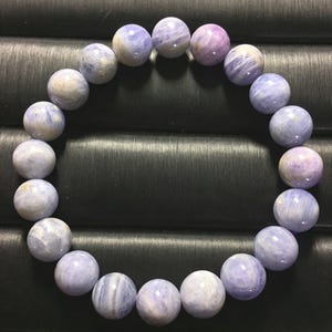 Könnte beinhalten: Ein kreisförmiges Stretch-Armband aus glatten, runden Perlen in den Farben Hellviolett, Lavendel und sanftem Blau. Jede Perle weist eine natürliche Marmorierung und durchscheinende Äderungen auf, die ein abwechslungsreiches Muster bilden.