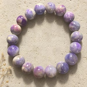 May include: A circular bracelet composed of smooth, polished spherical beads in varying shades of purple, lavender, and white. The beads feature a marbled, semi-translucent appearance with natural color variations throughout the stone surface.