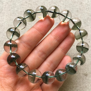 Op de afbeelding: Een cirkelvormige armband bestaande uit gladde, bolvormige kralen van helder kwarts met zichtbare groene fantoominclusies. Elke kraal heeft een duidelijke donkergroene, mosachtige formatie die in de transparante kristalstructuur is gevangen.