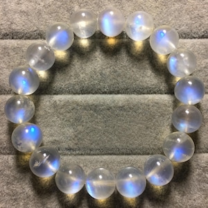 May include: A circular bracelet composed of smooth, translucent, spherical moonstone beads. Each bead exhibits a distinct, glowing blue adularescence, creating a luminous effect against the clear, polished surfaces of the round gemstone spheres.