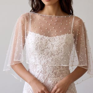 May include: A sheer, off-white tulle capelet featuring scattered white pearl embellishments. The capelet has a wide, boat-style neckline and drapes over the shoulders, worn over a white, sleeveless, floral-patterned lace dress with a straight neckline.