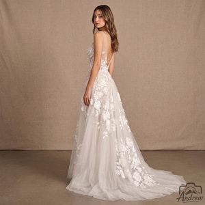 May include: A floor-length sleeveless gown in a light beige hue, featuring a sheer illusion neckline and back. The dress is adorned with white floral lace appliqués that cascade down the bodice and onto the full, layered tulle skirt.