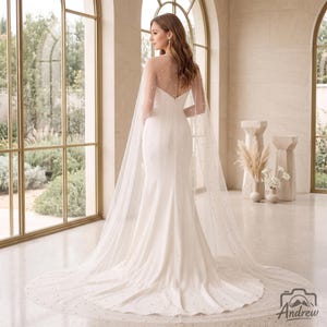 May include: A floor-length white gown with a fitted bodice and a flared skirt, featuring a sheer, floor-sweeping cape overlay adorned with scattered white pearls. The garment has a high, sheer neckline and a long, flowing train that extends outward.