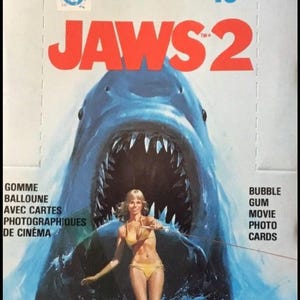 Może przedstawiać: Opakowanie gumy do żucia i kart ze zdjęciami z filmu Jaws 2 z dużym niebieskim rekinem z otwartą paszczą za osobą w żółtym bikini. Tekst zawiera Jaws 2, 15 cents, Pee Chee oraz Bubble Gum Movie Photo Cards z naklejką w każdym opakowaniu.