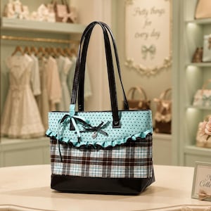 Op de afbeelding: Een tote bag met een zwarte onderkant, een bruin en blauw geruit middengedeelte en een lichtblauw bovenpaneel met zwarte stippen. De tas is voorzien van een ruche, twee zwarte schouderbanden en een decoratieve strik met een kleine bedel.
