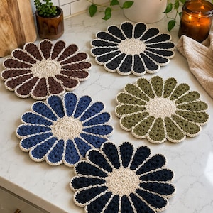 Peut inclure: Cinq dessous de verre circulaires en crochet en forme de marguerites, chacun doté d'un centre couleur crème et de pétales de couleurs unies distinctes, dont marron foncé, noir, vert olive, bleu marine et bleu clair, bordés de fil crème.