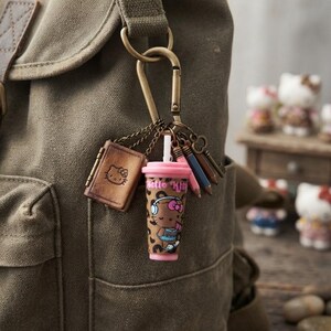 May include: A bronze-toned metal carabiner keychain attached to an olive green canvas backpack. The keychain features a small brown book charm with a Hello Kitty face, a pink and brown beverage cup charm with Hello Kitty text, and two miniature pencils.