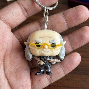 May include: A keychain featuring a small, stylized vinyl figure of an elderly character with gray hair, pointed ears, and yellow glasses. The figure wears a dark pinstriped suit, holds a small brown object in one hand, and an open book in the other.