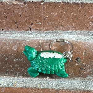 May include: A green turtle keychain featuring a textured shell with a white stripe along the center. The turtle is depicted in a side profile view with its head raised and is attached to a circular silver-toned metal split ring for keys.