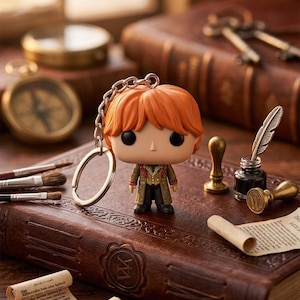 Pode incluir: Um porta-chaves com figura de vinil representando uma personagem com cabelo laranja, olhos pretos grandes e um casaco detalhado em tons de bege e verde escuro. A figura está presa a uma corrente e argola de metal prateado, em pé com nariz.