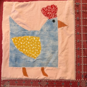 May include: A square fabric quilt block featuring a light blue chicken appliqué on a pale pink background. The chicken has a red patterned comb, a yellow floral wing, and an orange beak and feet. A black button serves as the eye. A ruler shows the block is 25.4 cm wide.