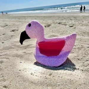 Peut inclure: Une peluche flamant rose avec un corps en tissu rose clair à motif floral, une aile en tissu rose foncé uni et un bec noir. Le flamant rose possède un œil noir unique visible et présente des coutures apparentes sur les bords du tissu.