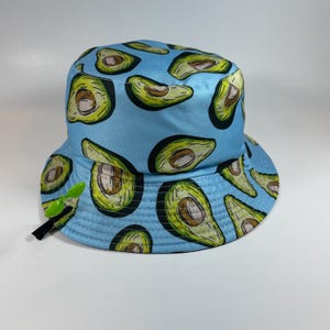 Op de afbeelding: Een lichtblauwe bucket hat met een herhalend patroon van doorgesneden groene avocado's met bruine pitten. De hoed heeft een brede, naar beneden lopende rand en een klein, felgroen plastic accessoire in de vorm van een spruitje.