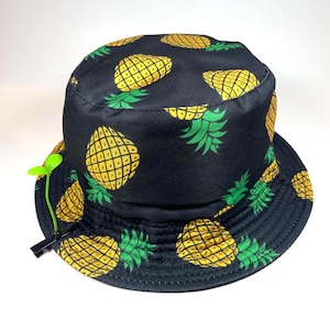 Può includere: Un cappello da pescatore nero con un motivo ripetuto di ananas gialli con foglie verdi. Un piccolo accessorio in plastica verde brillante a forma di germoglio è fissato sul lato della tesa con una clip nera, estendendosi verso l'alto dal bordo.