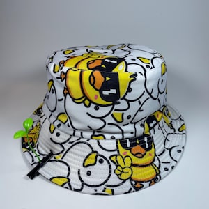 Op de afbeelding: Een witte bucket hat met een all-over print van gele cartoon-eenden met een zwarte zonnebril. Sommige eenden maken een vredesteken. Een kleine groene plastic clip in de vorm van een spruitje is aan de rand van de hoed bevestigd.