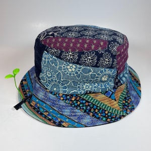 Op de afbeelding: Een patchwork bucket hat met diverse stofpatronen in blauw-, paars-, groen- en goudtinten. De kroon toont bloem- en geometrische prints, terwijl de rand diagonale strepen en een kleine groene plastic plantenspruit-clip als versiering heeft.