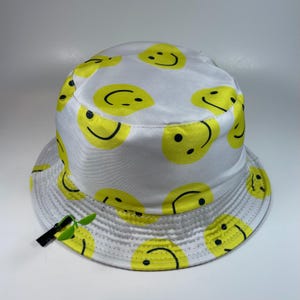 Op de afbeelding: Een witte bucket hat met een herhalend patroon van gele smileygezichten met zwarte ogen en gebogen monden. De hoed bevat een klein groen plastic plantje als accessoire dat met een zwarte clip aan de rand is bevestigd.