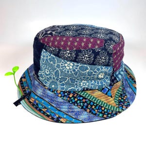 Puede incluir: Un sombrero de pescador de patchwork con diversos paneles de tela azul, morada y verde con variados patrones florales y geométricos. Un pequeño accesorio de plástico verde brillante en forma de brote está sujeto al ala con un clip negro.