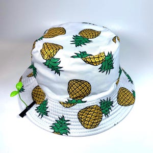 Può includere: Un cappello a secchiello bianco con un motivo ripetuto di ananas gialli con foglie verdi. Una piccola clip nera con un germoglio in plastica verde brillante è fissata sul lato del cappello, sporgendo oltre la tesa.