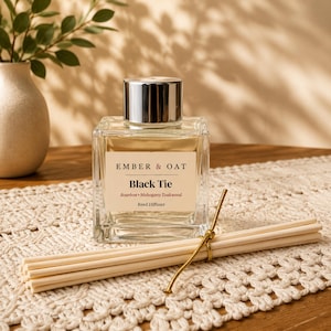 Könnte beinhalten: Ein quadratischer Glasflakon für einen Raumduft mit silbernem Verschluss und einem Etikett mit der Aufschrift Ember & Oat Black Tie Bourbon Mahogany Teakwood Reed Diffuser. Ein Bündel heller Holzstäbchen, gebunden mit einer goldenen Schnur, liegt davor.
