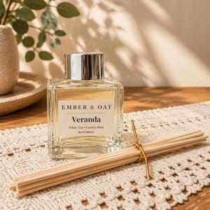 May include: A square glass reed diffuser bottle with a silver cap and a white label reading Ember & Oat Veranda White Tea Garden Mint. A bundle of light-colored wooden reeds tied with a thin gold cord rests in front of the glass container.