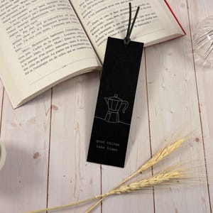 Pode incluir: Um marcador de página retangular preto com um desenho de linha branca de uma cafeteira moka e o texto "good things take time" impresso em branco. O marcador possui um laço de cordão preto preso através de um orifício no topo central.