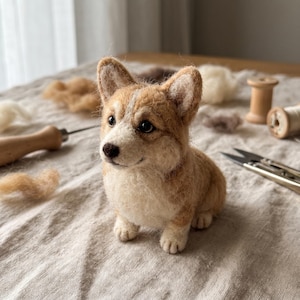 Könnte beinhalten: Eine nadelgefilzte Corgi-Figur aus beigefarbenen und weißen Wollfasern. Die kleine Hundeskulptur besitzt aufrecht stehende Ohren, dunkle Augen und eine dunkelbraune Nase, die die markanten körperlichen Merkmale der Rasse in Textur darstellen.
