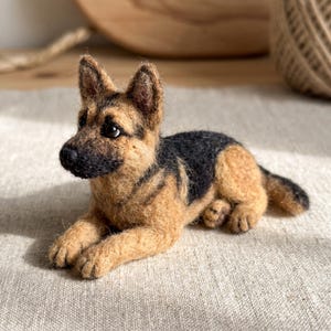 May include: A needle-felted German Shepherd dog figurine in a lying position. The sculpture features a black muzzle, dark ears, and a coat blending tan and black wool fibres. It has small, dark, glossy eyes and a textured, soft appearance.