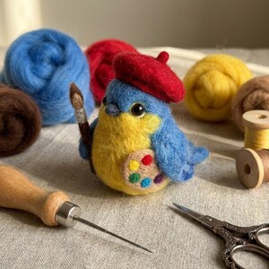 May include: A needle-felted blue and yellow bird figurine wearing a small red beret. The bird holds a miniature paintbrush and features a felted artist palette with colorful dots on its side, surrounded by wool roving and crafting tools.