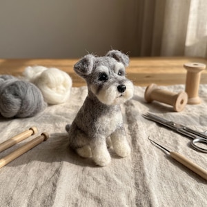 May include: A needle-felted miniature Schnauzer dog figurine with grey and white wool fur, black eyes, and a black nose. The small sculpture is positioned among crafting supplies including wooden spools, metal scissors, and wool roving.