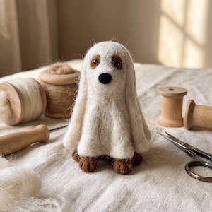 Puede incluir: Una figura de perro de lana fieltrada con un disfraz de fantasma blanco. El perro tiene orejas marrones, ojos oscuros y una nariz negra. El disfraz de fantasma está hecho de fibras de lana blanca, cubriendo el cuerpo y mostrando patas marrones.