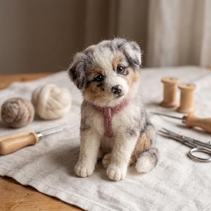 May include: A needle-felted puppy figurine with mottled gray, white, and tan fur, featuring dark eyes, a black nose, and a soft pink collar. The puppy is crafted with a textured, wool-like appearance and sits in an upright, alert posture.