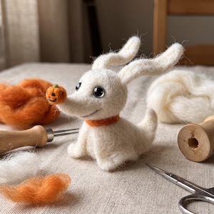 Puede incluir: Una figura de perro fantasma de lana fieltrada en blanco con orejas largas, ojos negros y un collar naranja. Una pequeña calabaza de fieltro naranja con una cara cosida a mano está equilibrada en la punta de su nariz de fibras de lana suaves.