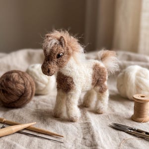 May include: A needle-felted miniature horse figurine with a white and brown coat, a fuzzy mane, and a tail. The horse has dark, glossy eyes and a textured, woolly appearance. It stands upright, showcasing detailed craftsmanship in its soft fiber construction.