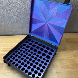 May include: A square storage case with a hinged lid featuring a purple and blue iridescent geometric star pattern. The interior base is black with a grid of circular cutouts, also showing a purple and blue iridescent finish on the surface.